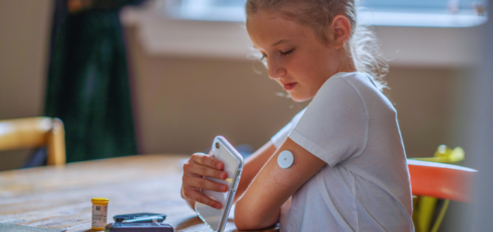 Dziewczynka z cukrzycą typu 1 mierzy poziom glukozy za pomocą telefonu i sensora umieszczonego na ramieniu. Siedzi przy stole, obok leżą akcesoria diabetologiczne.