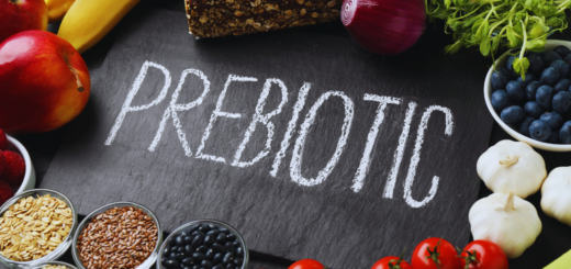Czarny prostokątny blat z napisem „PREBIOTIC” zapisanym białą kredą. Wokół rozłożone są produkty bogate w błonnik i naturalne prebiotyki, takie jak jabłka, banany, cebula, czosnek, borówki, maliny, pomidorki koktajlowe, zboża, siemię lniane, czarna fasola, chleb pełnoziarnisty i świeże zioła.