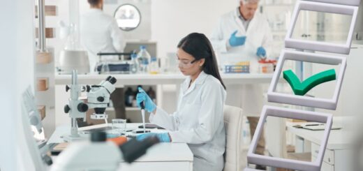 W laboratorium badawczym kobieta w białym fartuchu i rękawiczkach wykonuje analizę przy mikroskopie, a w tle pracują inni naukowcy. Na obrazie widoczny jest zielony symbol odhaczenia, symbolizujący rzetelność i potwierdzoną jakoś badań.
